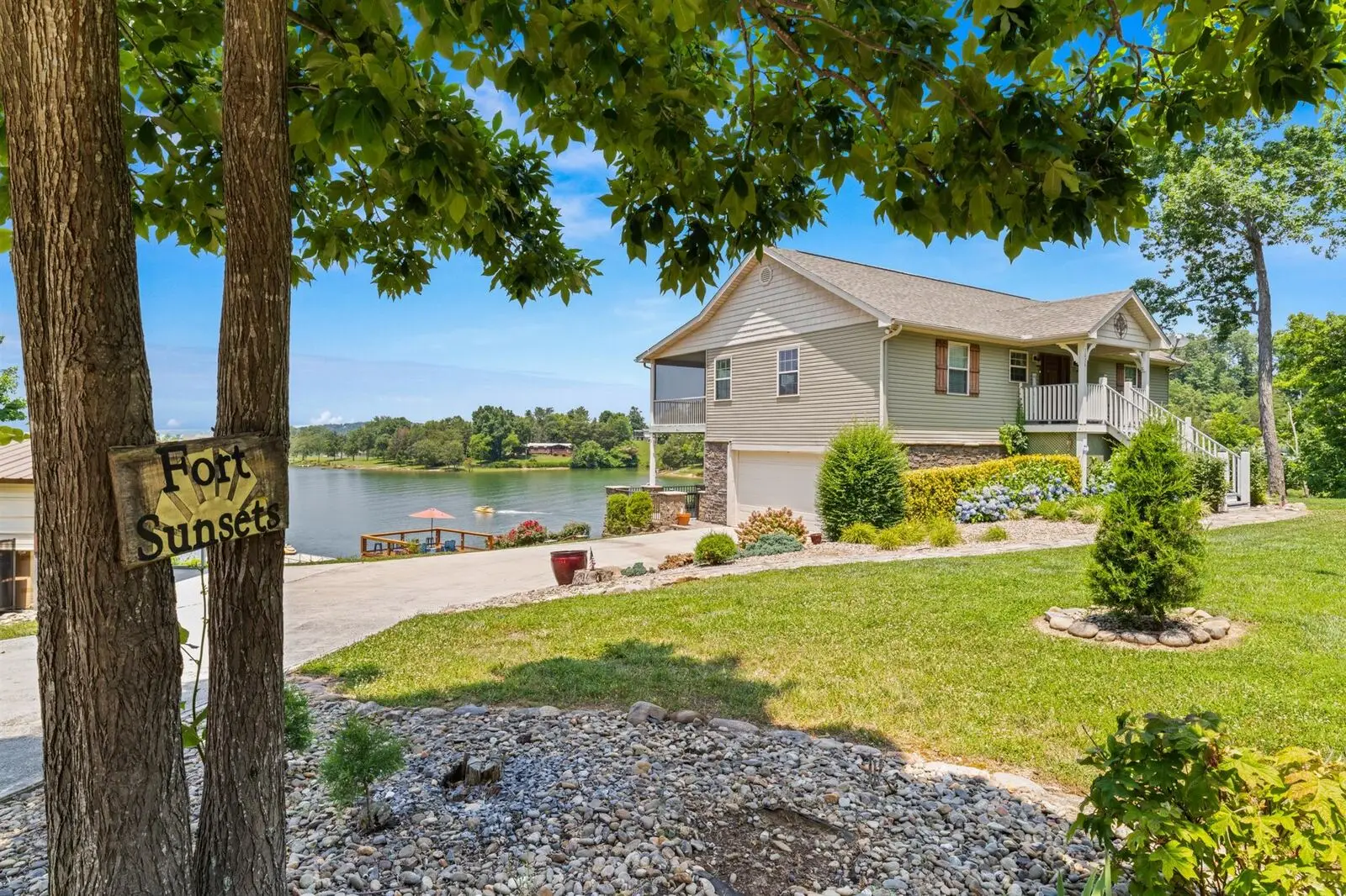 Front view of Fort Sunsets lakehouse with wooden Fort Sunsets sign on tree, landscaped yard with hydrangeas, Douglas Lake and floating dock visible in background