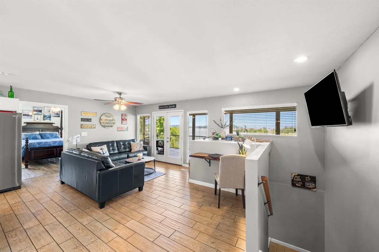 Main level living area with dark leather sectional sofa, ceiling fan, wall-mounted TV, lake-themed wall decor, french doors opening to the screened porch with Douglas Lake views, and glimpse of king bedroom to the left