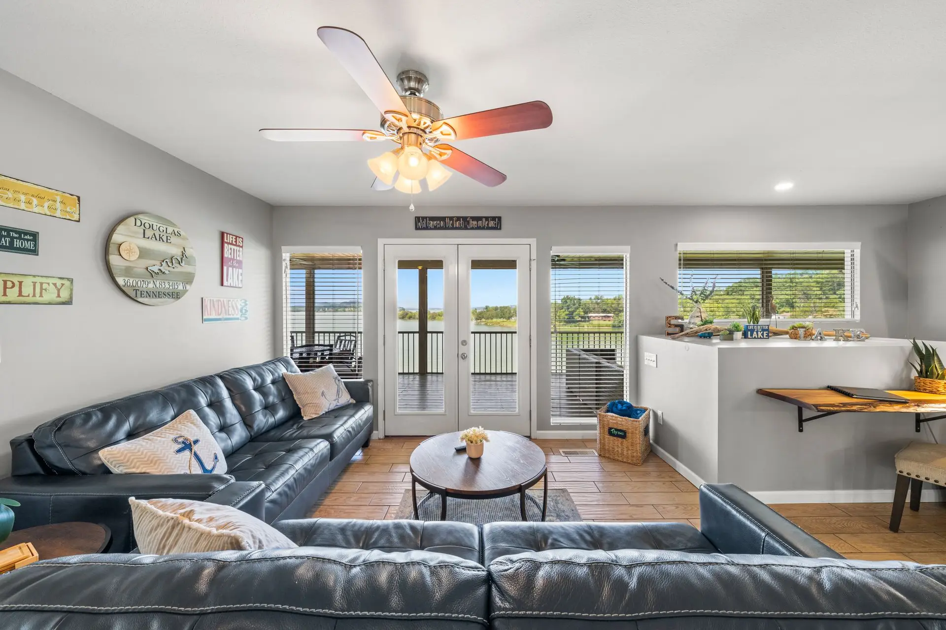 Living room seating area with large dark leather tufted sectional sofa, round wood and metal coffee table, Douglas Lake themed wall art, ceiling fan, and french doors with lake and hill views through the glass