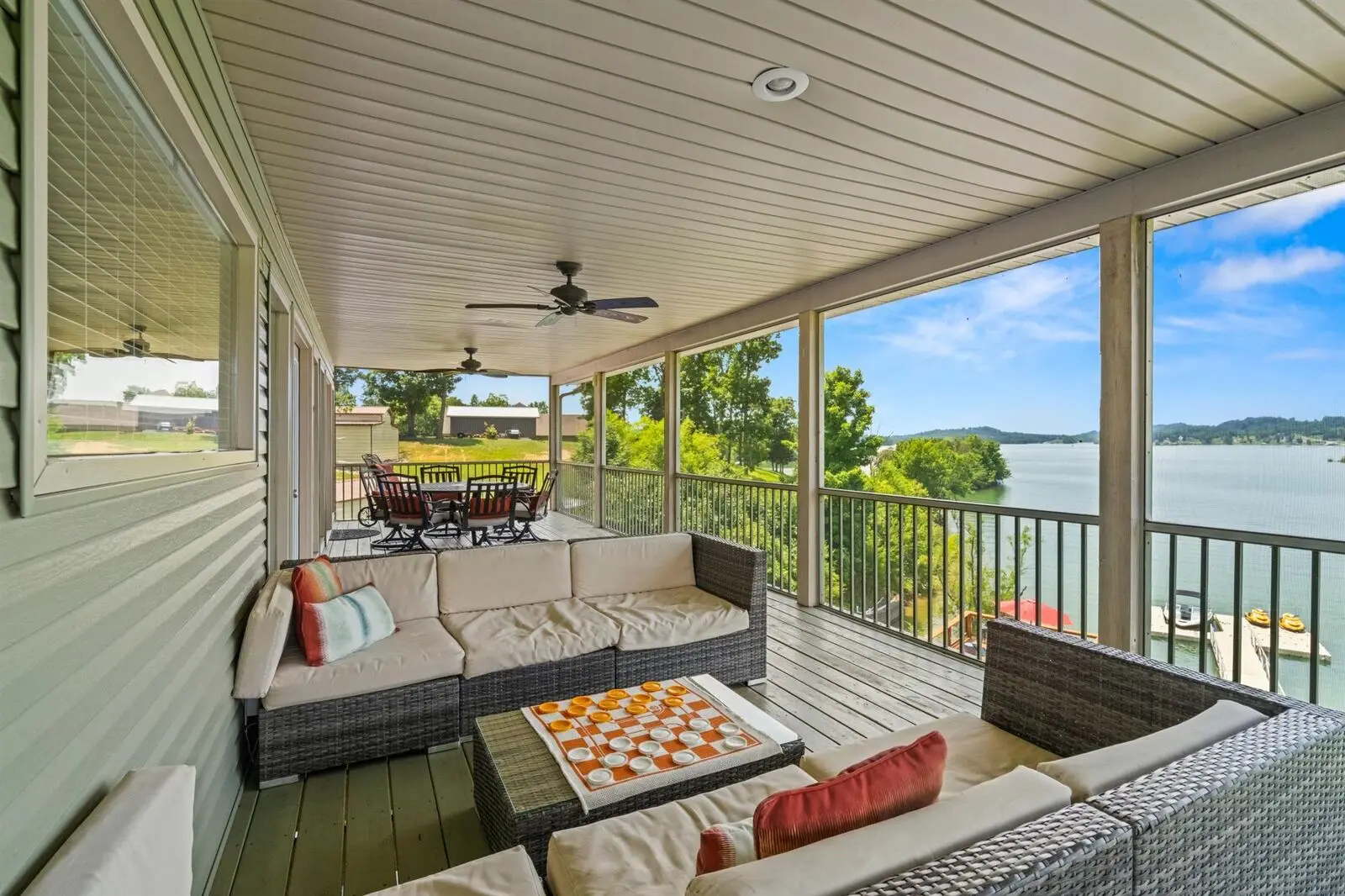 Spacious screened-in porch with wicker sectional sofa with cream cushions, checkerboard game on coffee table, dining area behind, and panoramic views of Douglas Lake and surrounding hills