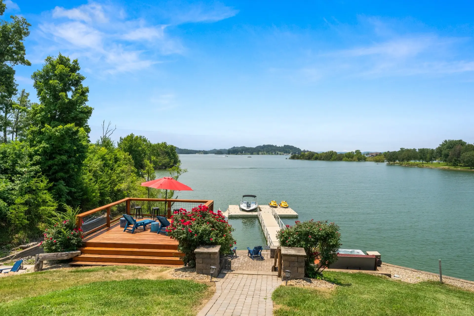 View from the house down to the lakeside area showing the wooden sun deck with blue Adirondack chairs and red umbrella, brick paver walkway with rose bushes leading to the floating dock with boat and jet skis, and hot tub to the right