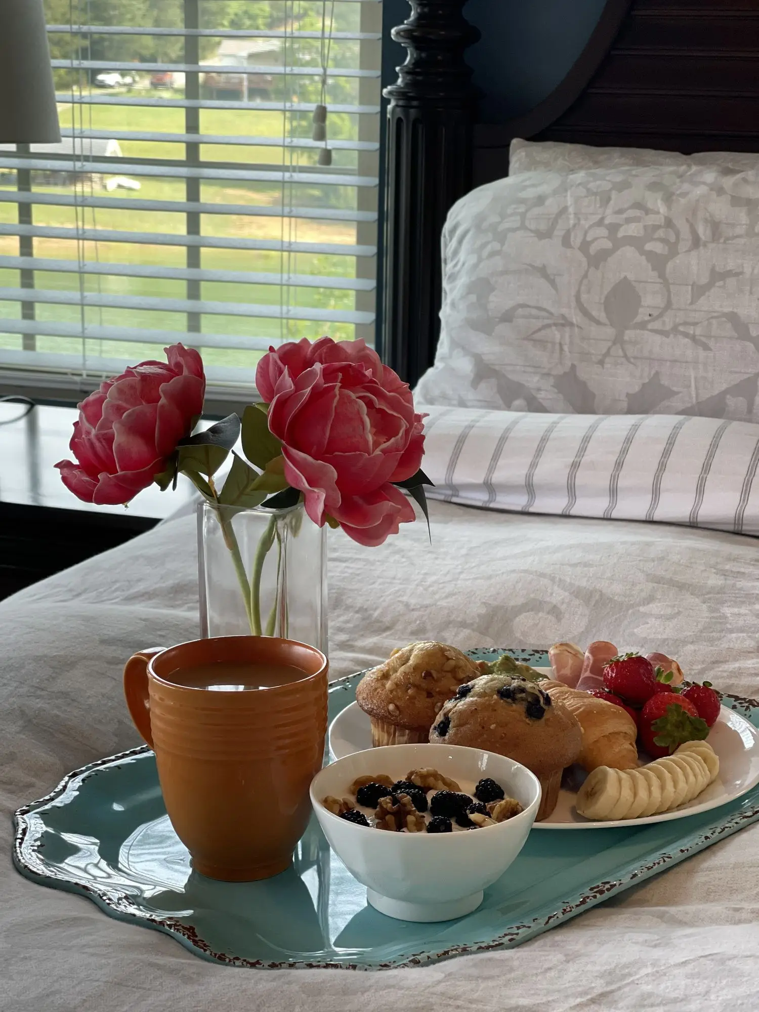 Breakfast tray with muffins, berries, coffee and pink peonies on the bed beside a lake-view window