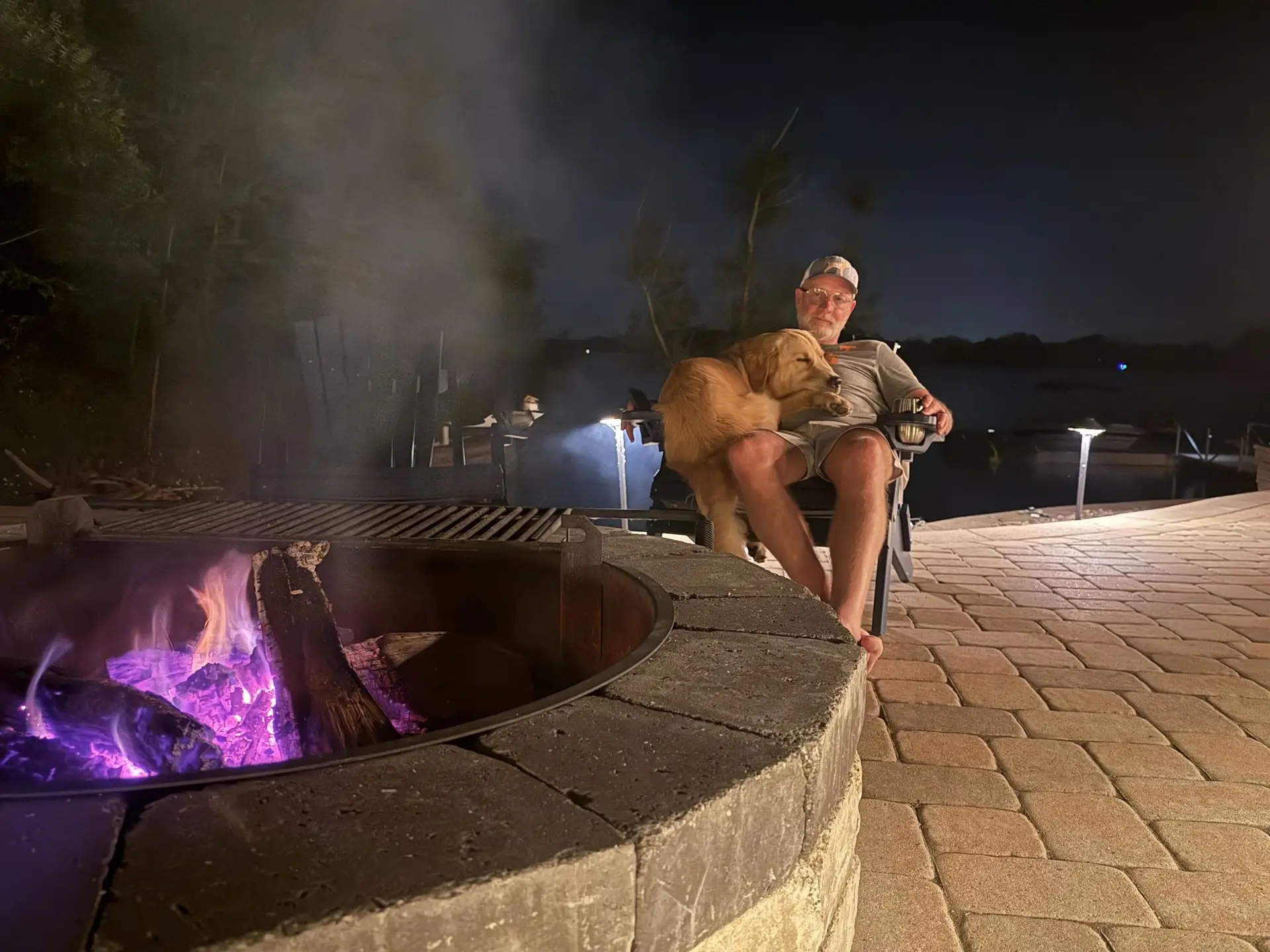 Purple and blue flames in the sunken fire pit at night with a man and his golden retriever beside it