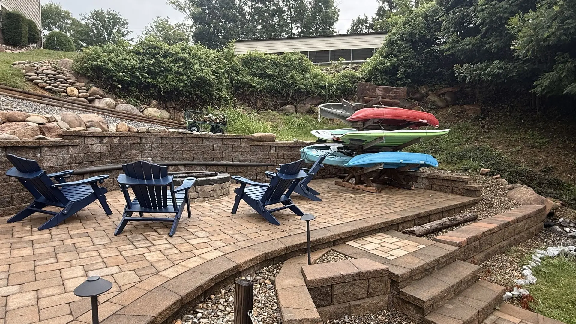 The sunken fire pit patio by day, Adirondack chairs circled and the kayak rack loaded behind