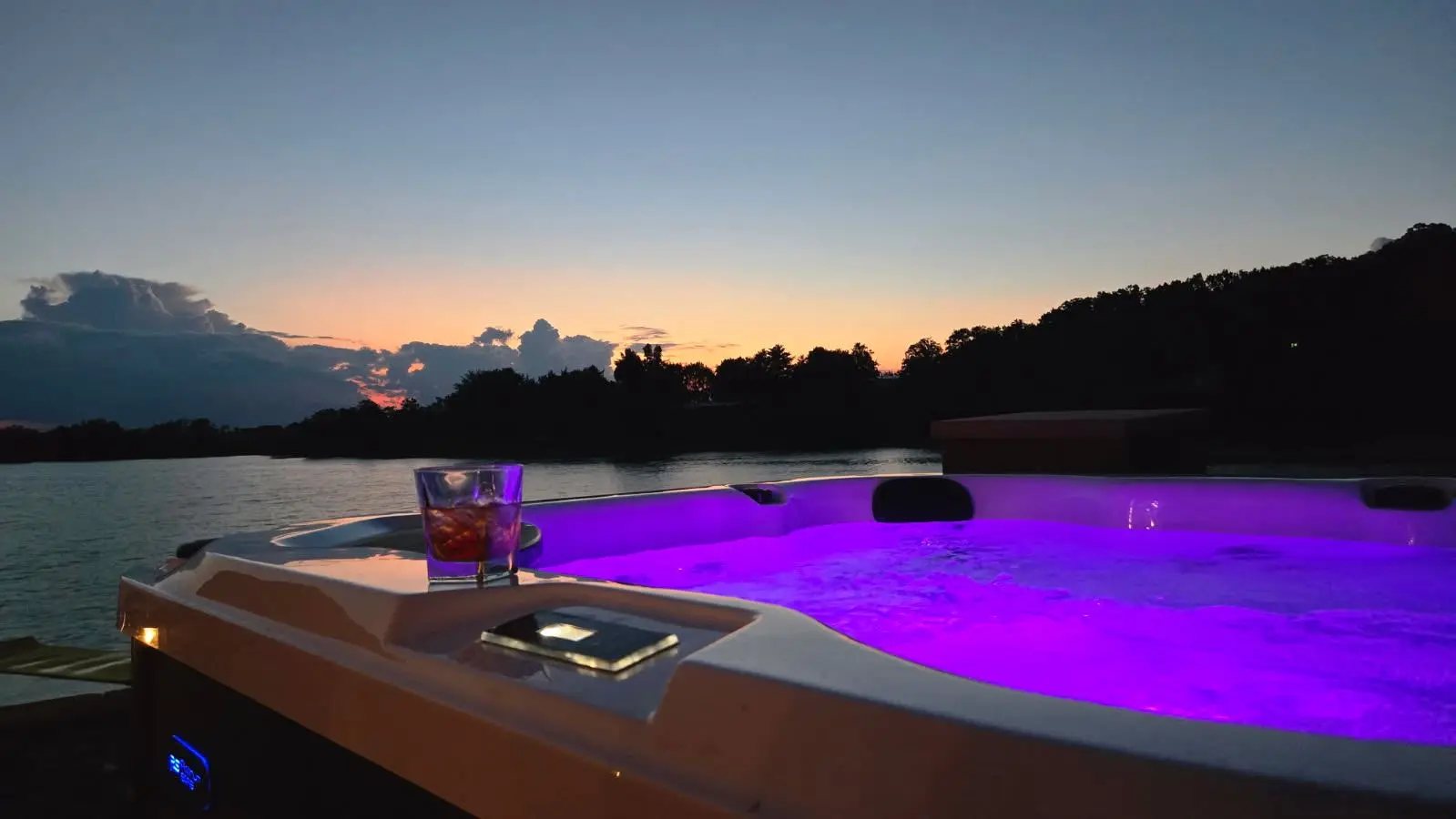 Hot tub glowing purple at dusk with drinks on the rim and the lake beyond